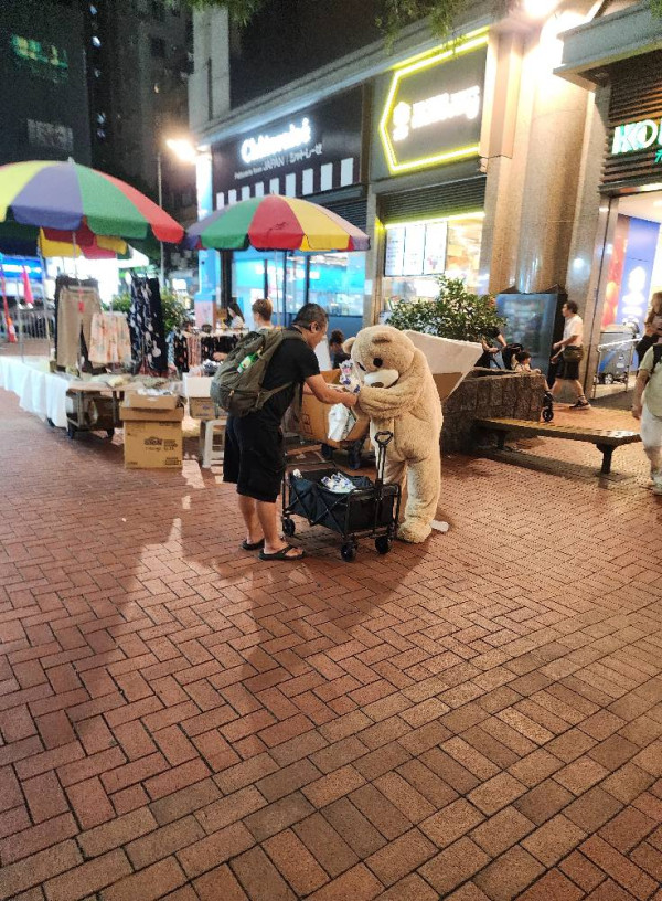 從照片見到，一名身穿啡色小熊布偶裝的人，正在街頭上和途人兜售鮮花，旁邊則放了一架手推車，可看到裡面放了不少鮮花，其中有一名中年男子主動上前買花，而身穿小熊布偶裝的人非常感謝，雙手奉上鮮花時更微微鞠躬。圖片來源：Facebook@全港店舖消息(開張/執笠)關注組