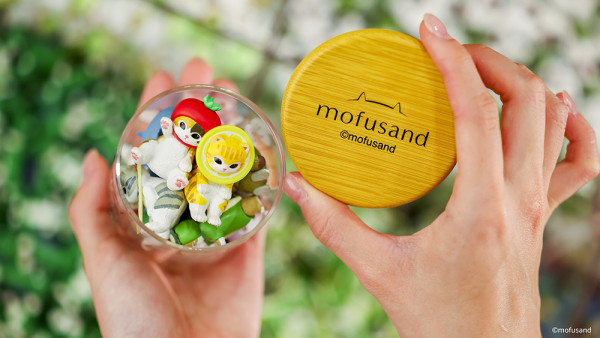 雙手拿着裝滿多款草莓與水果造型mofusand粒粒迷你公仔的透明玻璃樽，蓋子印有mofusand標誌 
