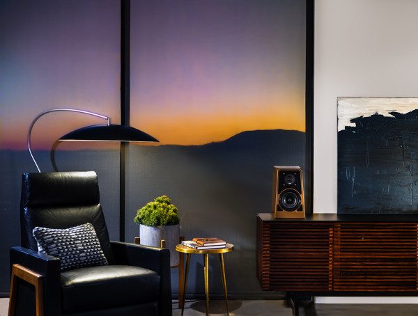 Mira Place的HiFi Gallery精选多款顶级音响设备设备，店面设计成具质感的居家角落，包含黑色皮质单人沙发、简约落地灯、木质音响柜，柜上放有 HiFi 书架喇叭与抽象画作，背景为夕阳景色。