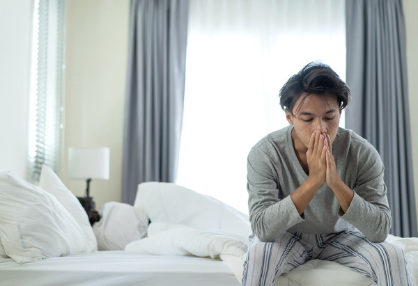 相片中一名亞洲男子身穿灰色長袖睡衣和條紋睡褲,坐在白色的床上。他雙手交疊掩著口鼻,低著頭,眼睛閉著或看向下方,顯得疲倦、沮喪或有些身體不適。床上有一名女子在睡覺,背景是臥室的窗戶和灰色的窗簾。