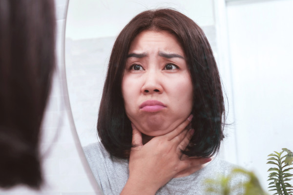 一名亞洲女性早晨照鏡子捏著下巴與面部浮腫肌肉，露出煩惱困惑的表情。